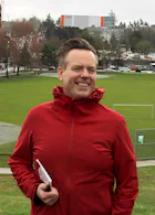Mark Wiens stands wearing a red jacket in front of a park and smiles at the camera